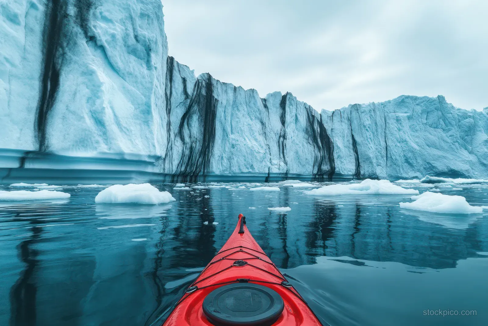 A red kayak moves across blue water toward icebergs beneath towering Antarctic glacier cliffs.