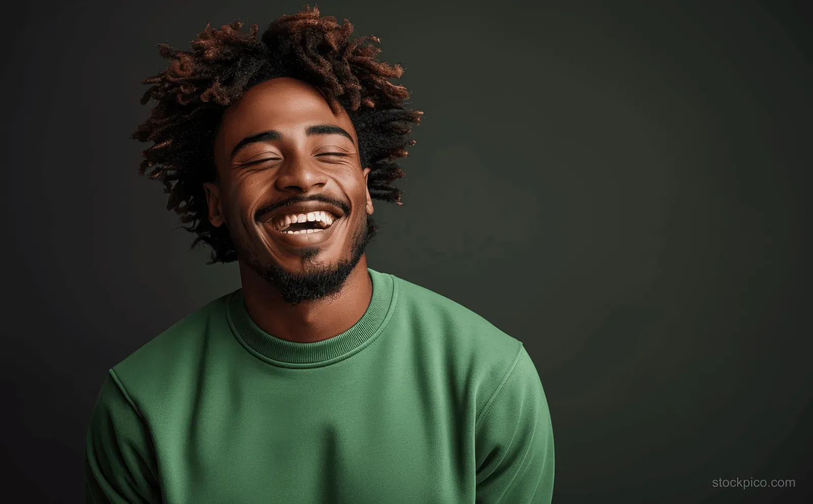 Young man in a green sweatshirt laughing in front of a dark studio background.