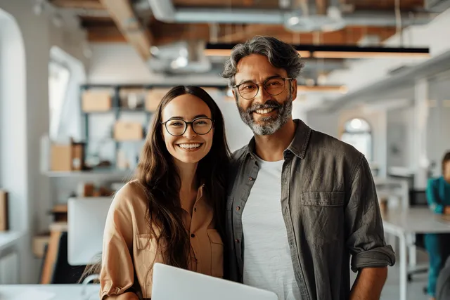 Smiling diverse coworkers pose with laptop in modern office hallway under warm light