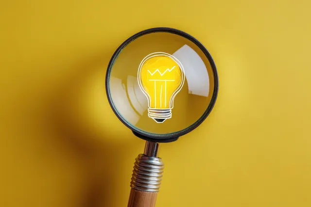 Magnifying glass enlarging a glowing light bulb icon over a bright yellow background.