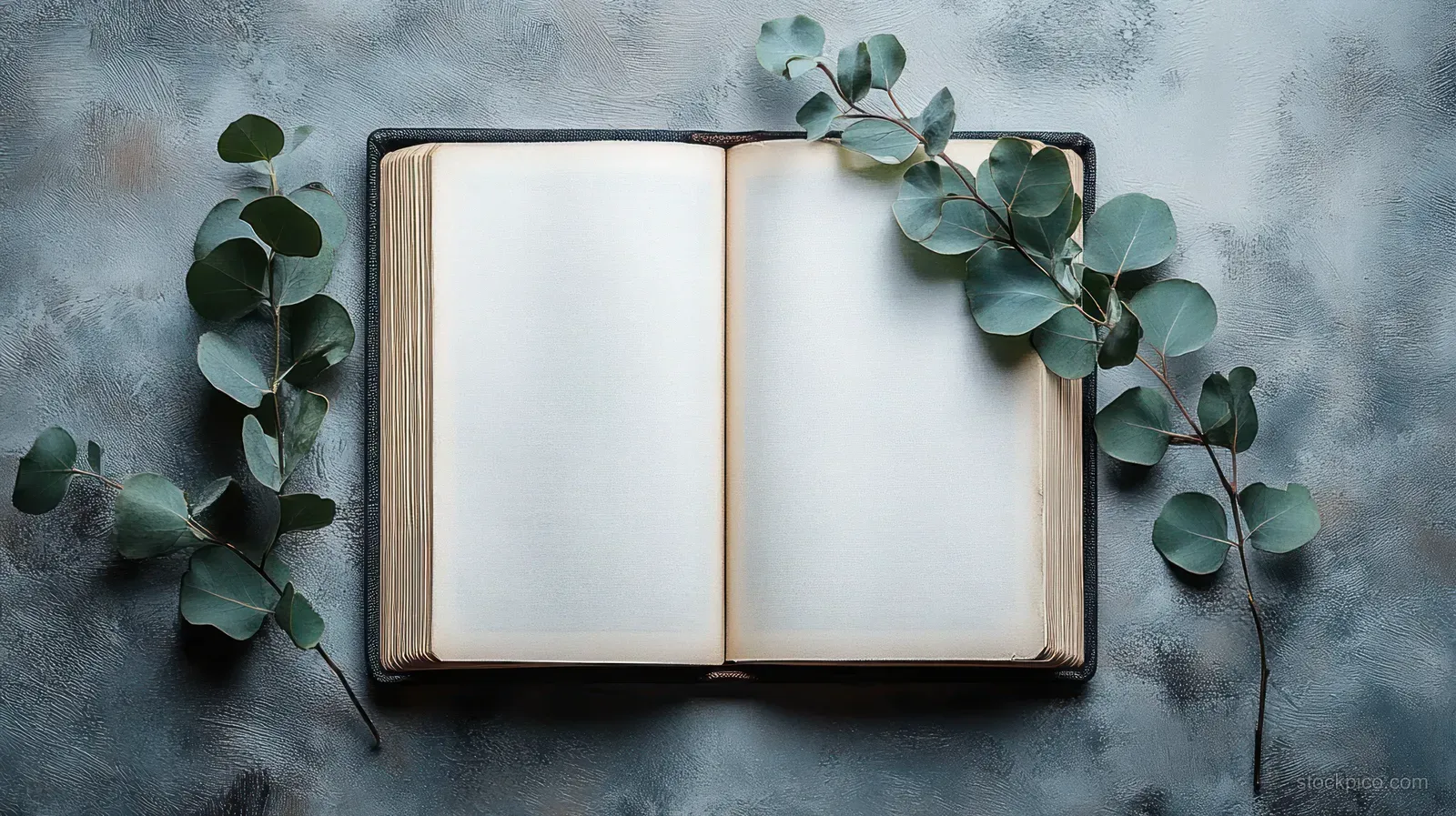 Open vintage blank book on a textured surface with eucalyptus leaves scattered around it.