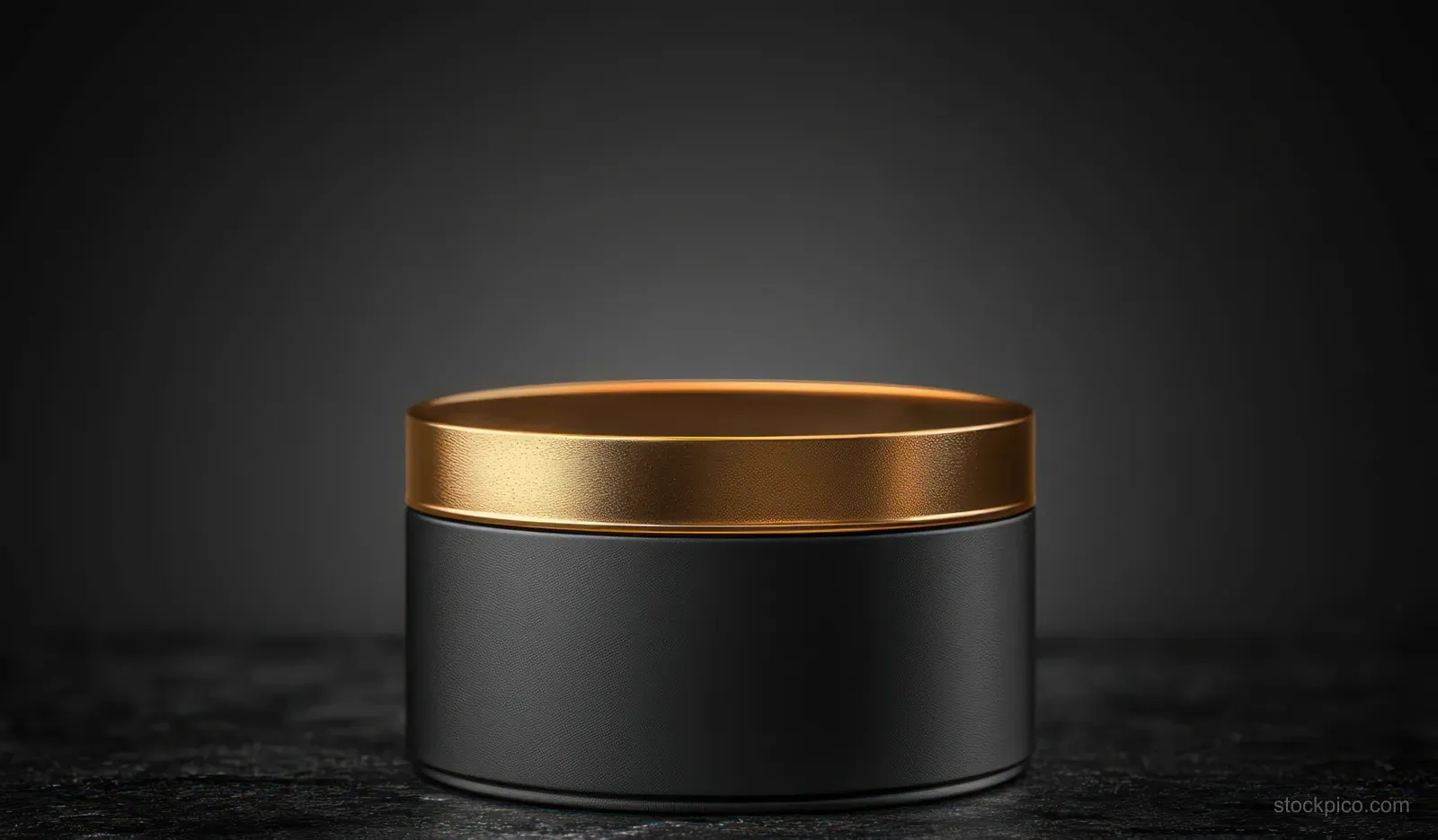 Matte black cosmetic jar with a reflective gold lid sits on a dark studio surface under soft spotlight glow.