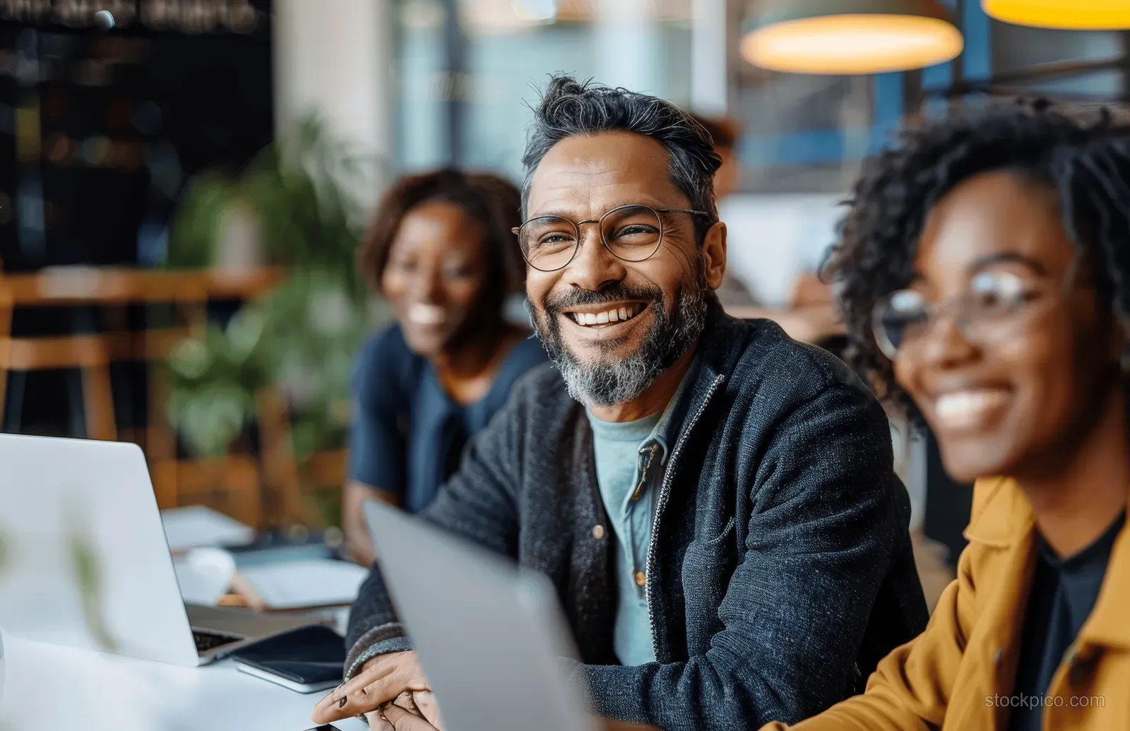 Multiethnic business professionals smile while collaborating with laptops during an office meeting in a bright room.
