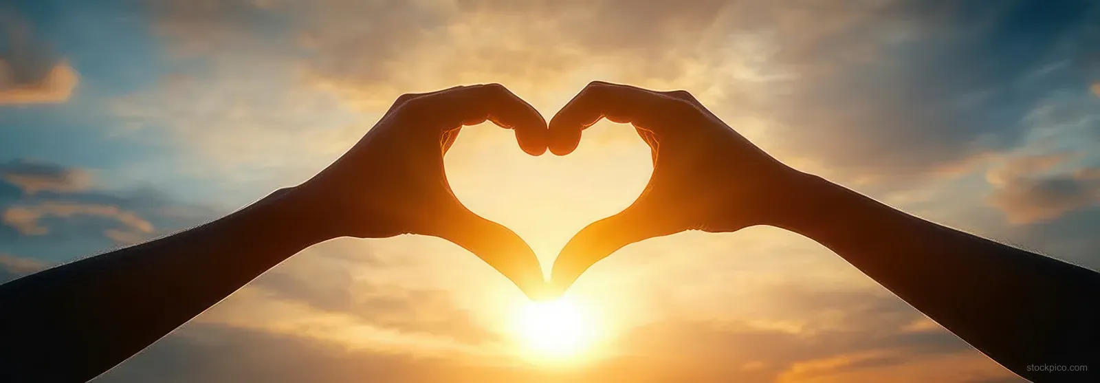Silhouetted hands form a heart around the glowing sun at sunset against a colorful clouded sky.