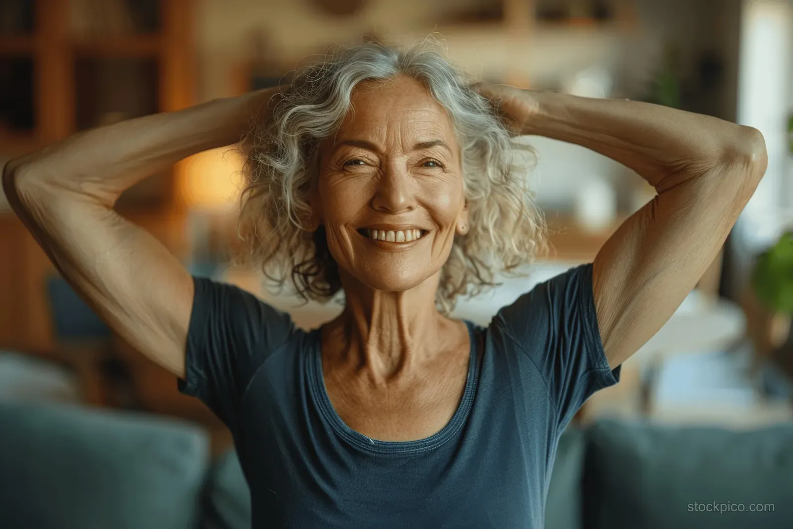 Smiling senior woman holds an at-home yoga stretch with arms behind head and a green sofa in view.